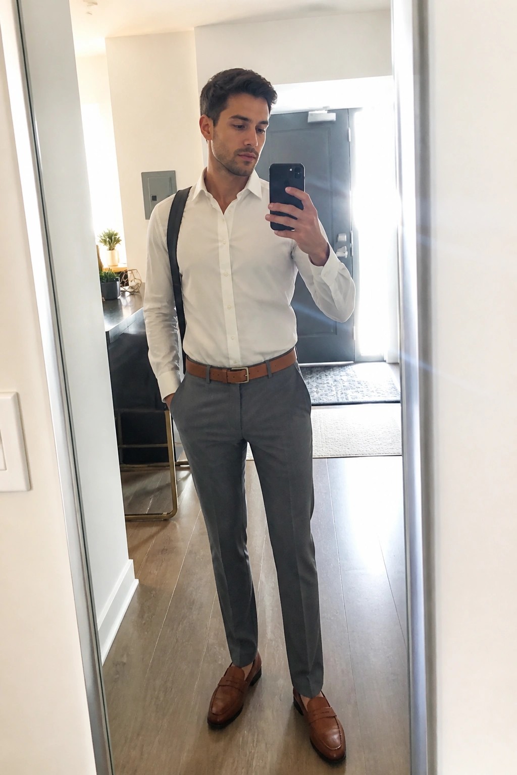 Man in white shirt and gray pants taking mirror selfie with phone.