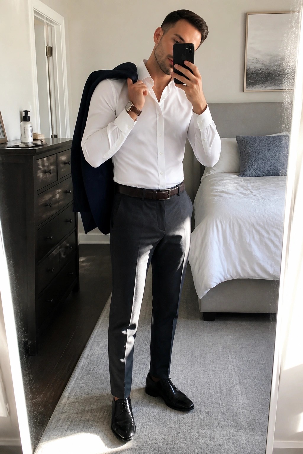 Man in white shirt and gray pants taking mirror selfie with navy blazer over shoulder