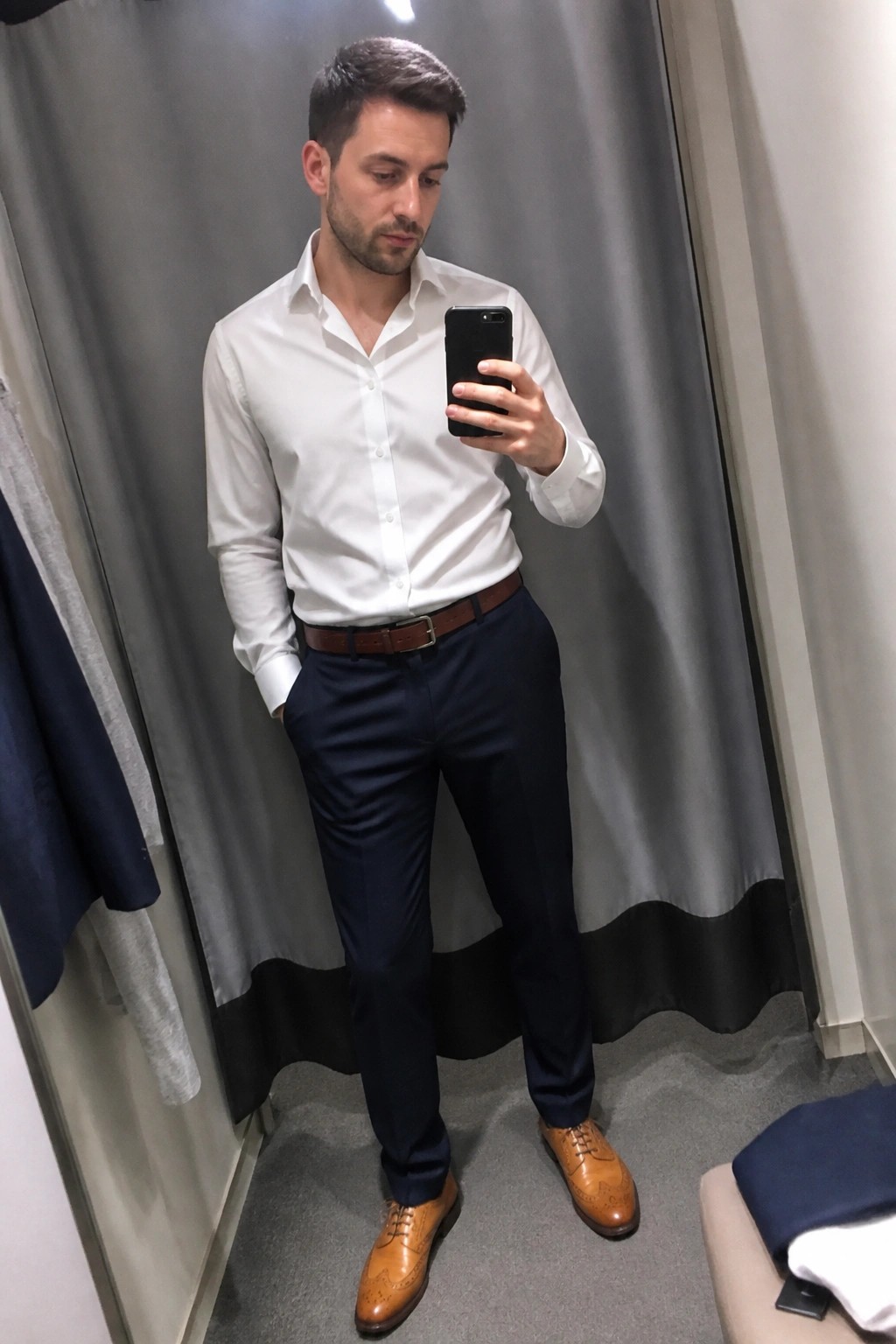 Man in white shirt and navy pants taking mirror selfie in fitting room with brown shoes.