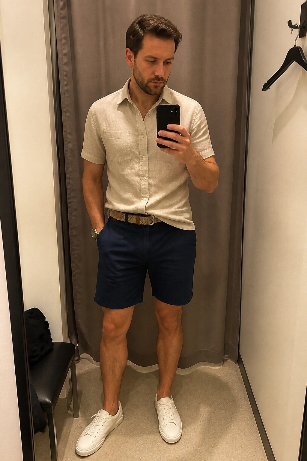 Man in beige shirt and navy shorts taking mirror selfie in fitting room