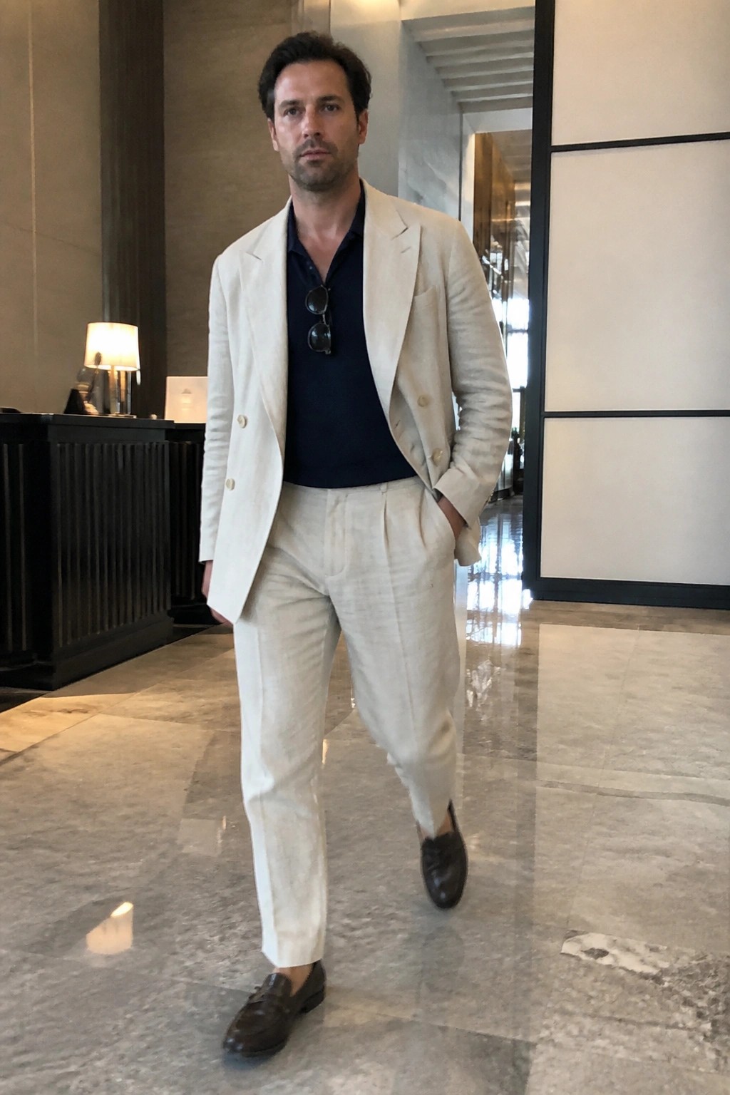 Man in beige linen suit and navy shirt stands in marble lobby with hand in pocket