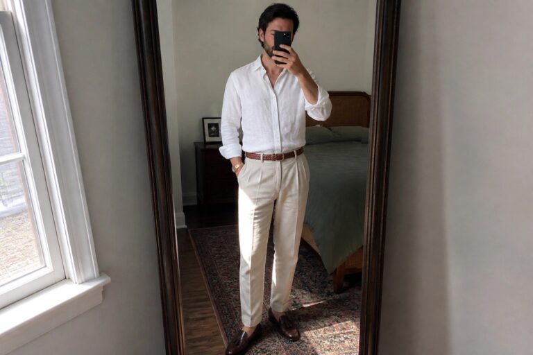 Man in white linen shirt and cream trousers taking mirror selfie in bedroom.