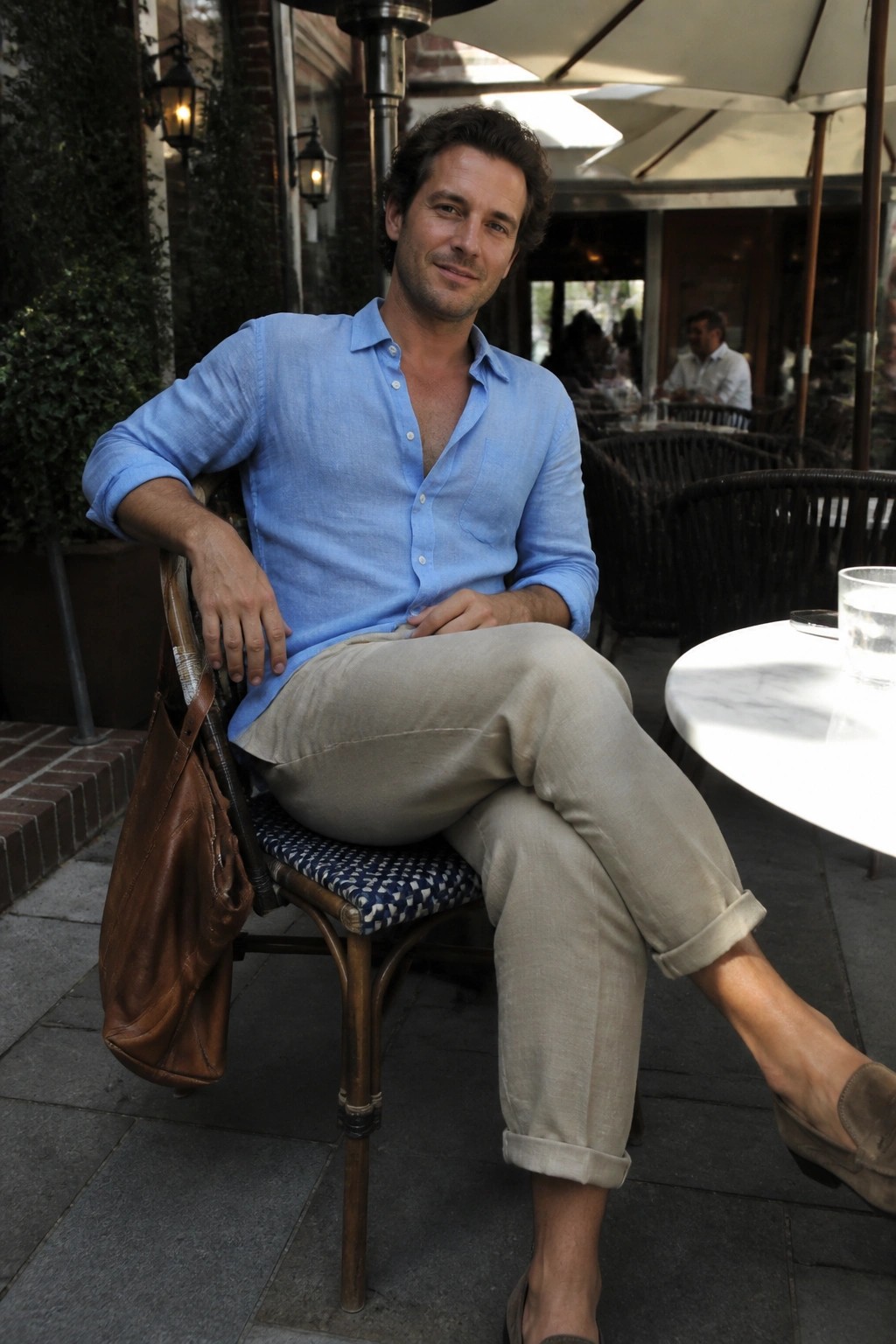 Man in blue shirt and beige pants at outdoor cafe with leather bag
