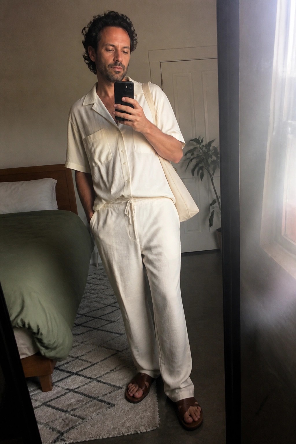 Man in cream linen shirt and pants taking mirror selfie in bedroom