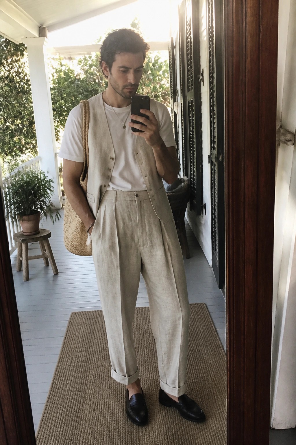 Man in beige linen vest and pleated trousers taking a selfie on porch.
