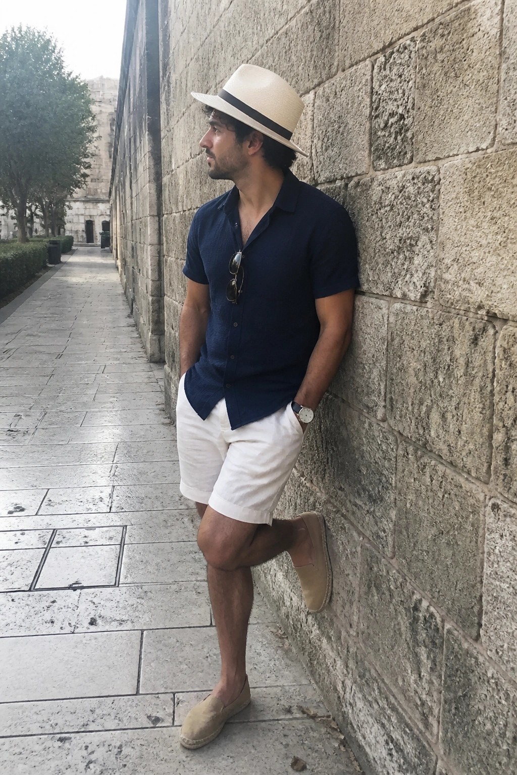 Man in navy shirt and white shorts leans against stone wall wearing hat