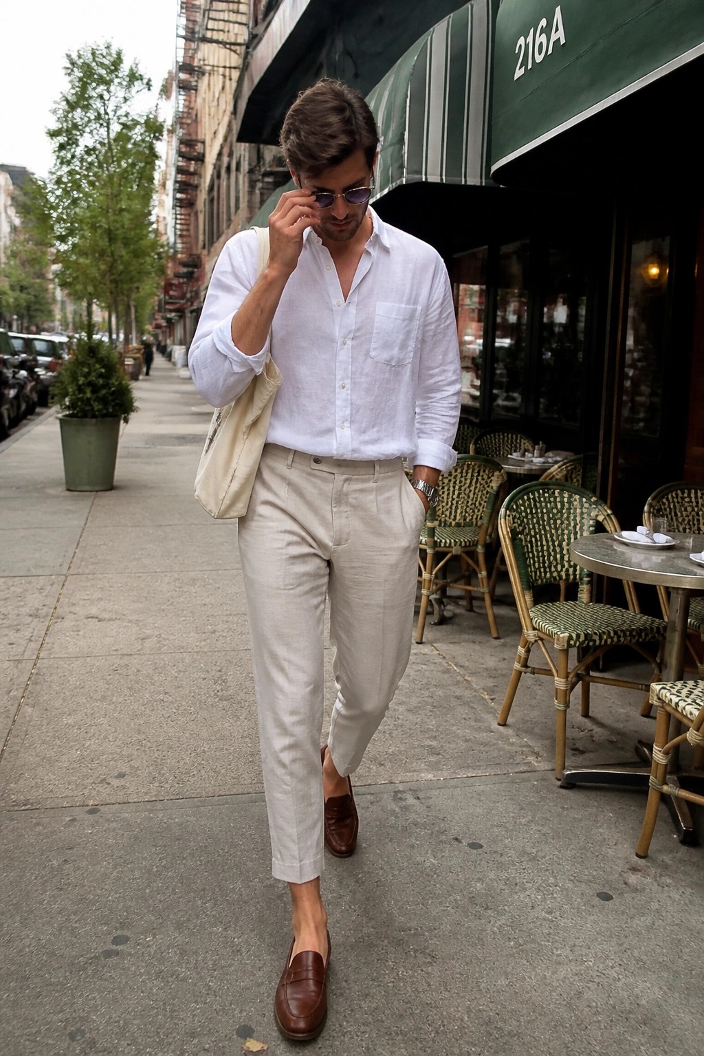 Man in white shirt and beige pants adjusts sunglasses on city sidewalk by cafe