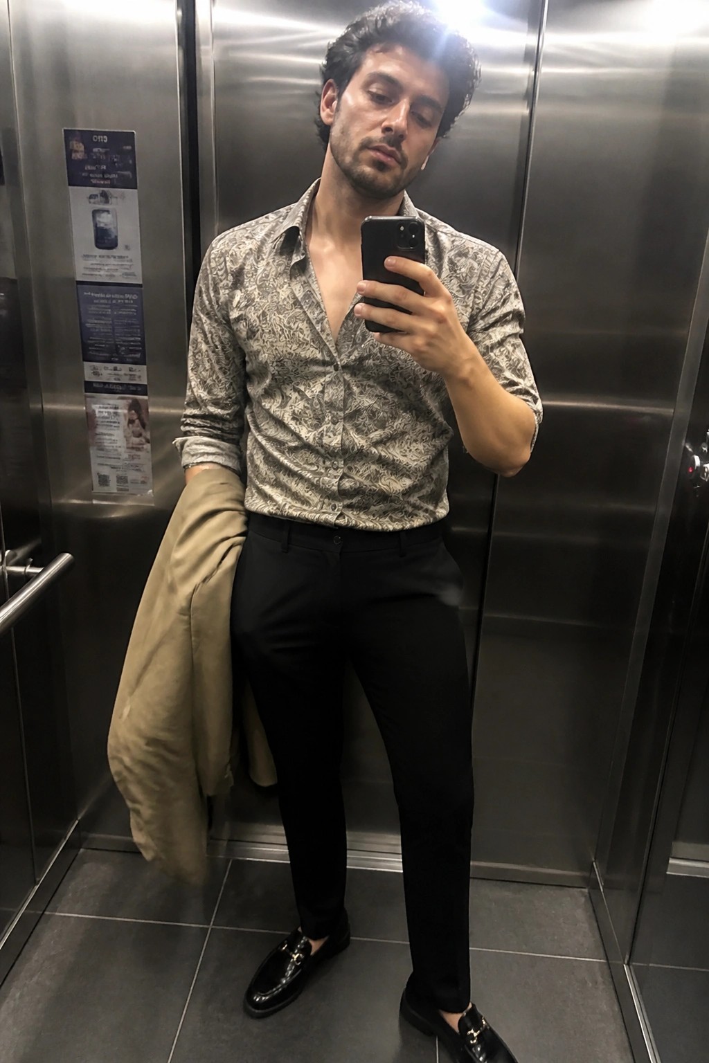 Man in patterned shirt takes selfie in elevator holding phone and jacket