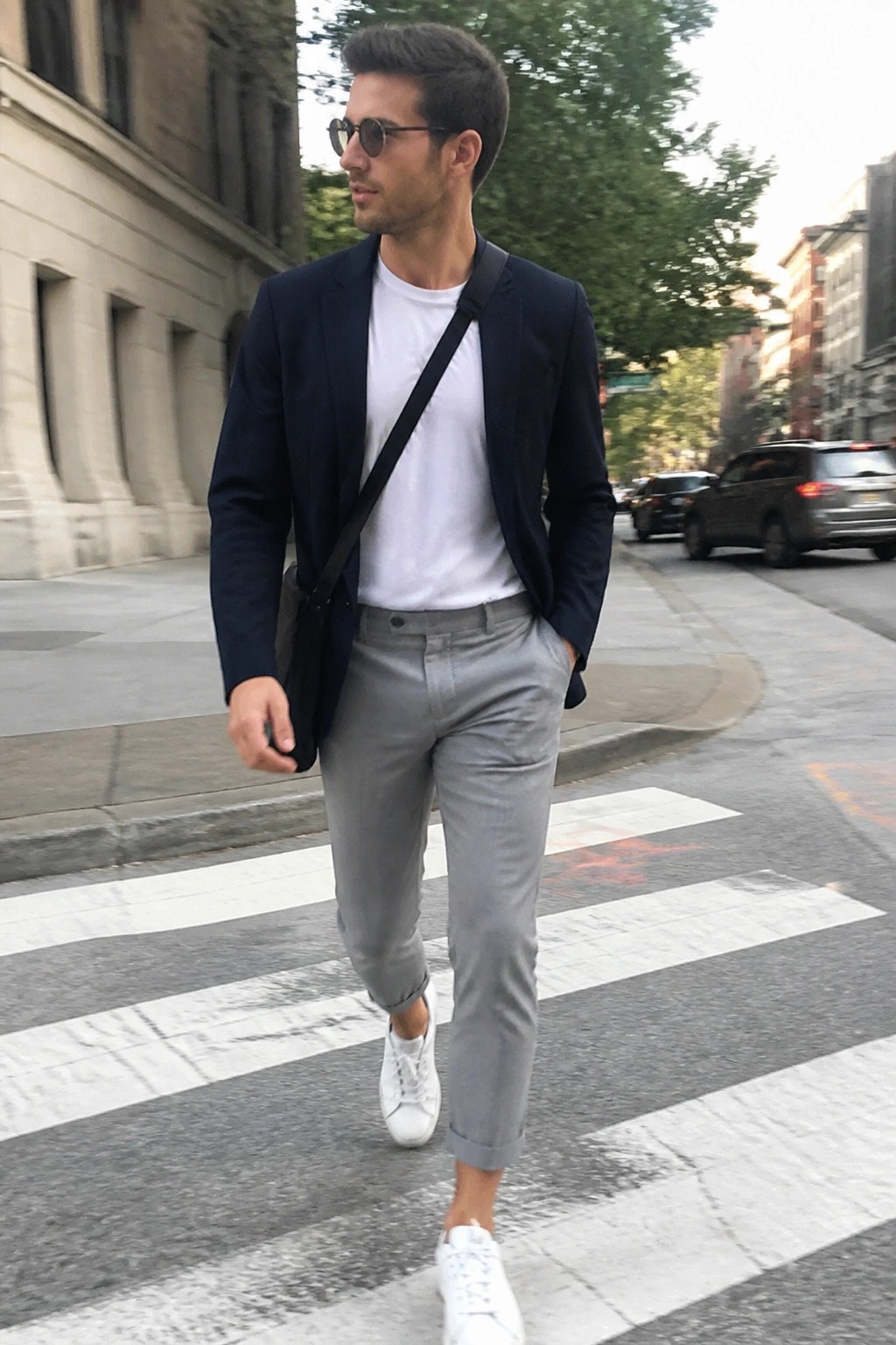 Man in navy blazer, white tee, and gray pants crossing urban street with bag.