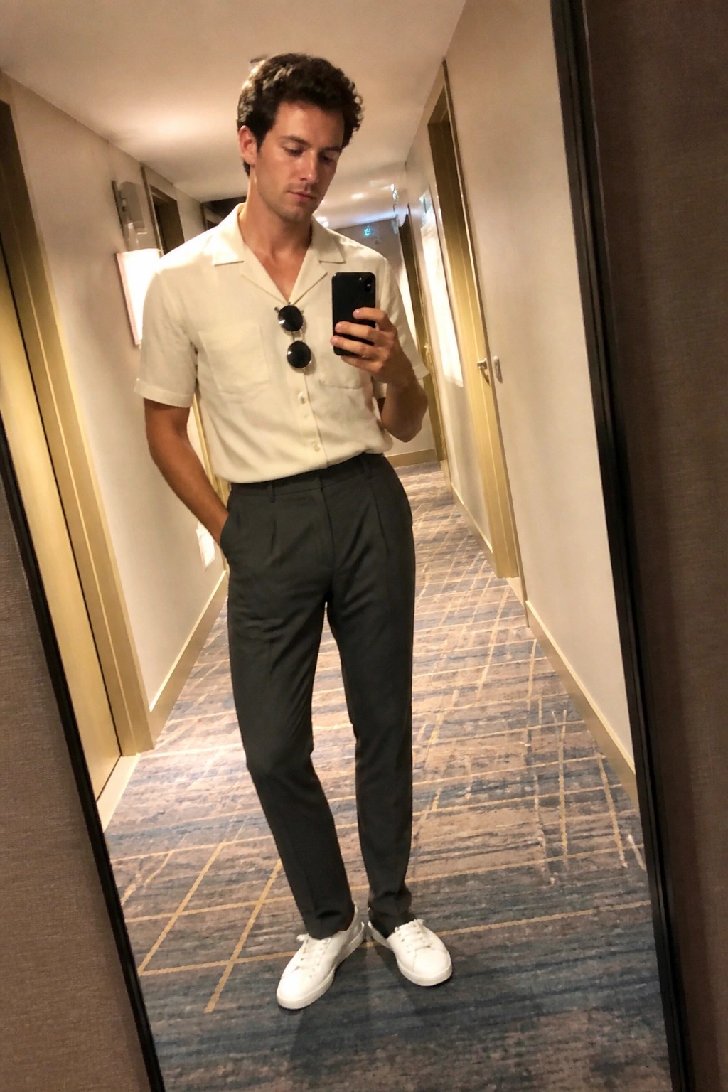 Man in a mirror selfie wearing dark gray pleated trousers, cream short sleeve button up shirt with sunglasses on the placket, and white sneakers in a hotel hallway.