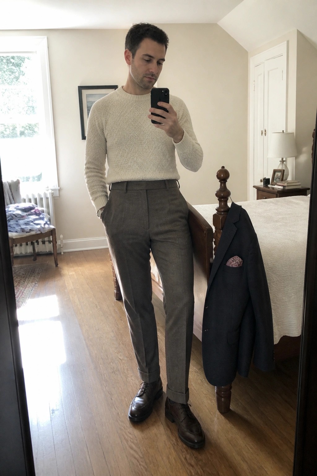 Man in cream sweater and gray trousers taking mirror selfie in bedroom