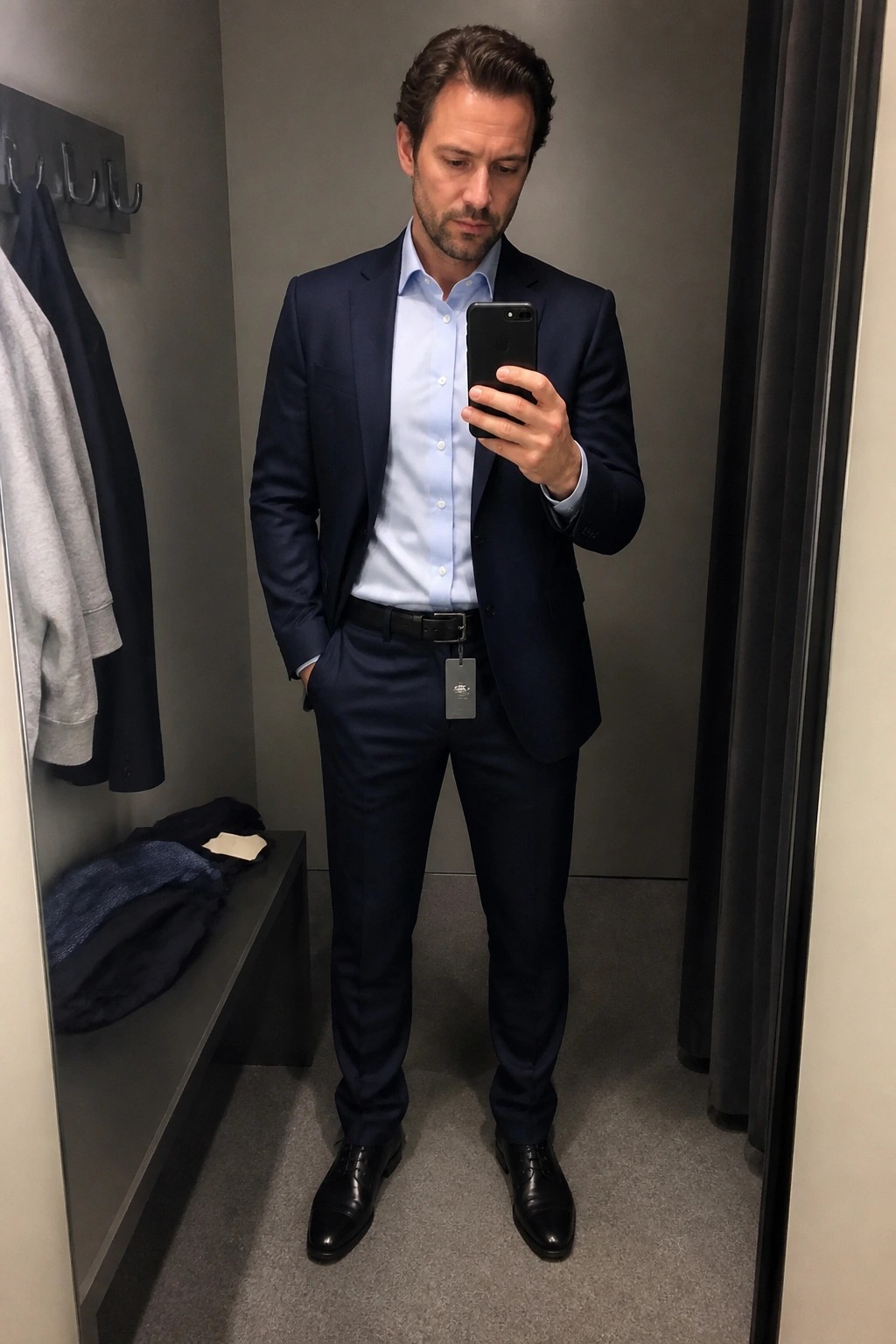 Man in navy suit and light blue shirt taking mirror selfie in fitting room.