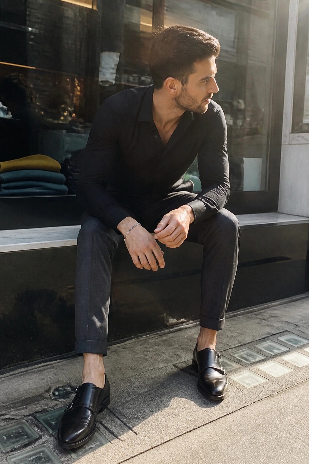 Bearded man in black shirt and monk shoes seated on urban ledge in sunlight.
