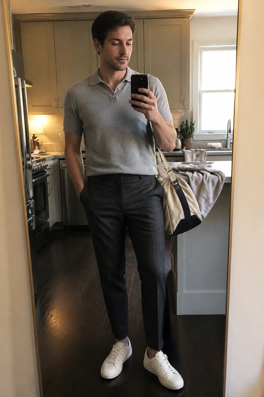 Man in gray polo shirt and dark pants takes mirror selfie in kitchen with bag