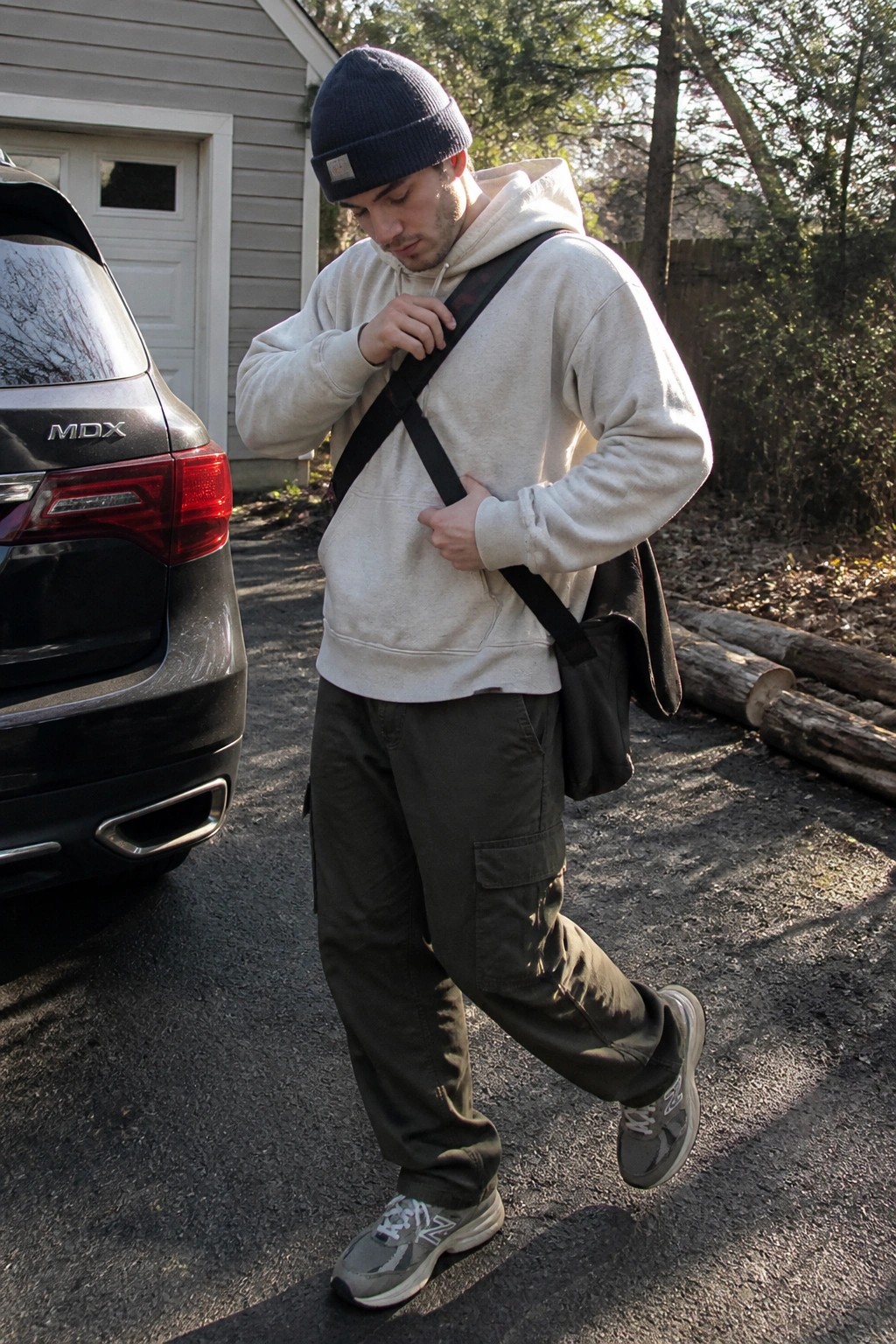 Man in cream hoodie and beanie adjusting black crossbody bag by SUV
