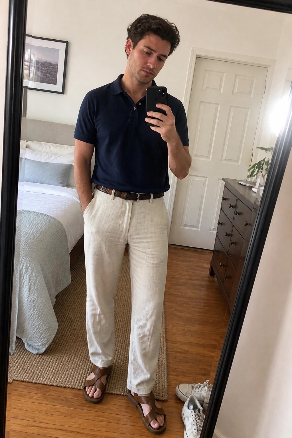 Man in a navy polo shirt tucked into relaxed off-white linen pants with a brown woven belt and brown leather sandals, shown in a full-length mirror selfie indoors.
