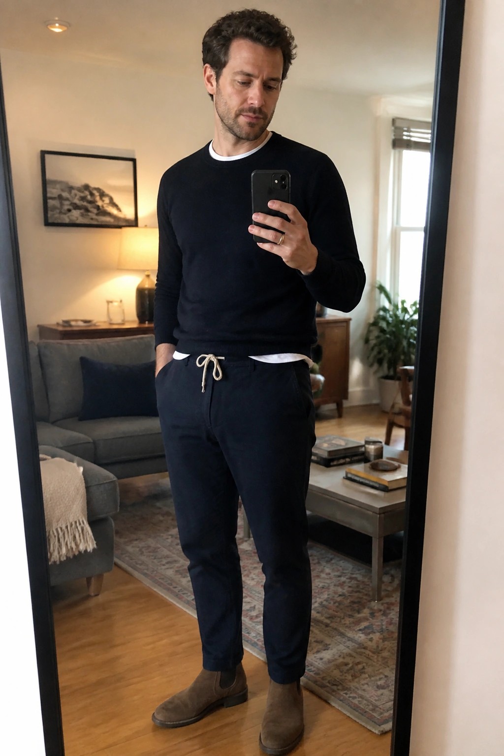 Man wearing relaxed navy pants with white drawstring, black crewneck sweater over white t-shirt, and brown suede chelsea boots while taking a mirror selfie.
