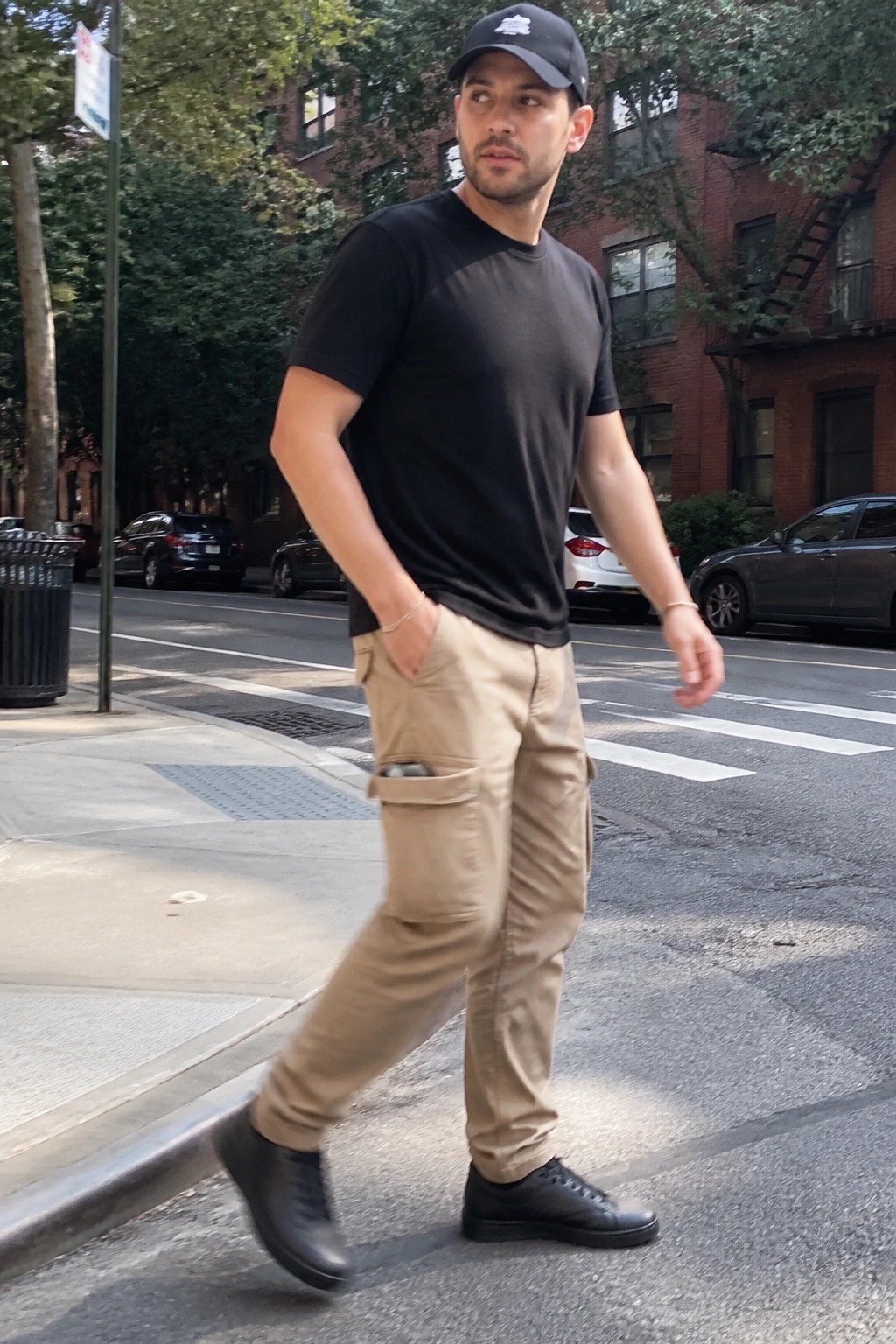 Man wearing relaxed beige cargo pants, black short sleeve tee, black cap, and black sneakers standing on a city sidewalk.