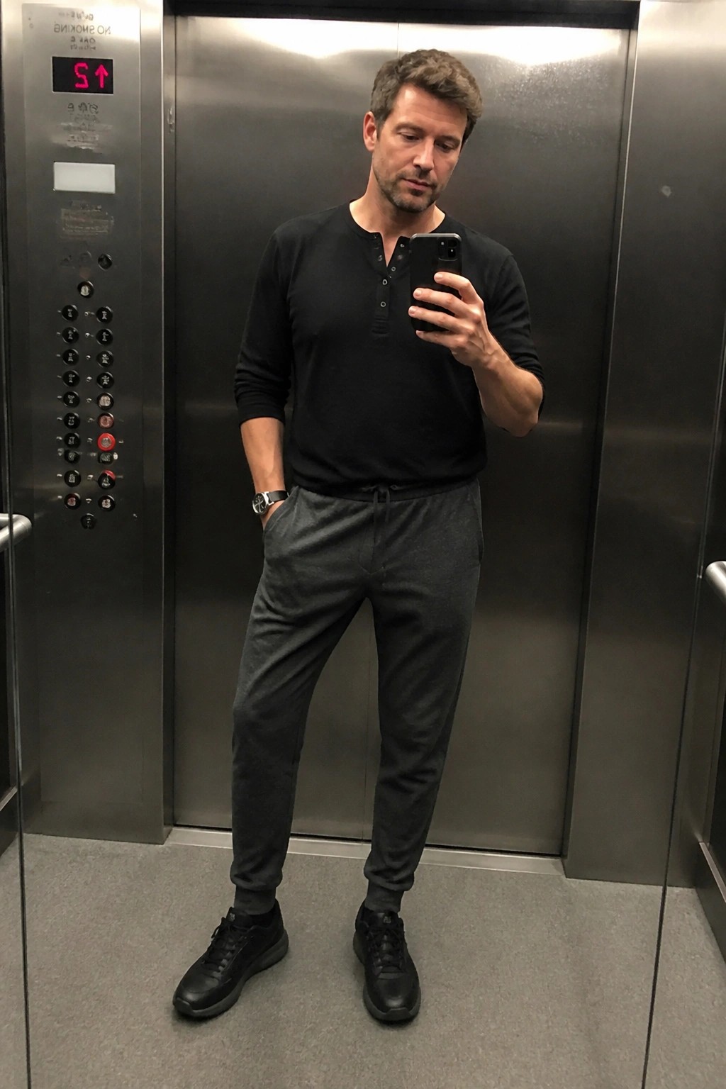 Man in black shirt and gray sweatpants taking selfie in elevator.