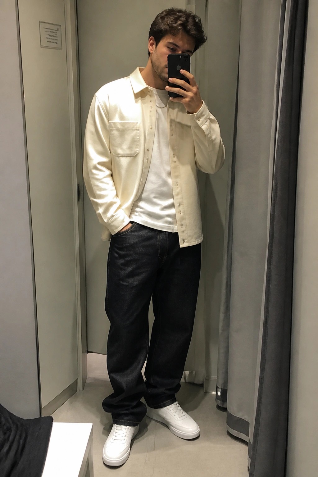 Man in cream shirt and dark jeans taking mirror selfie with phone