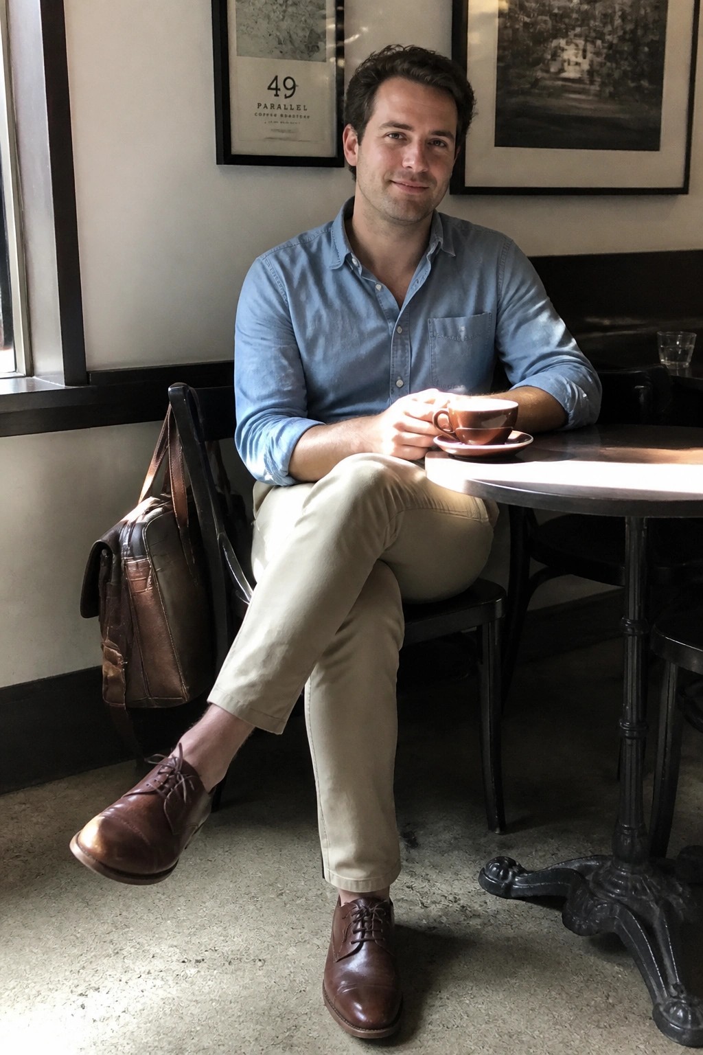 Man in blue shirt and khaki pants holding coffee beside leather bag in cafe