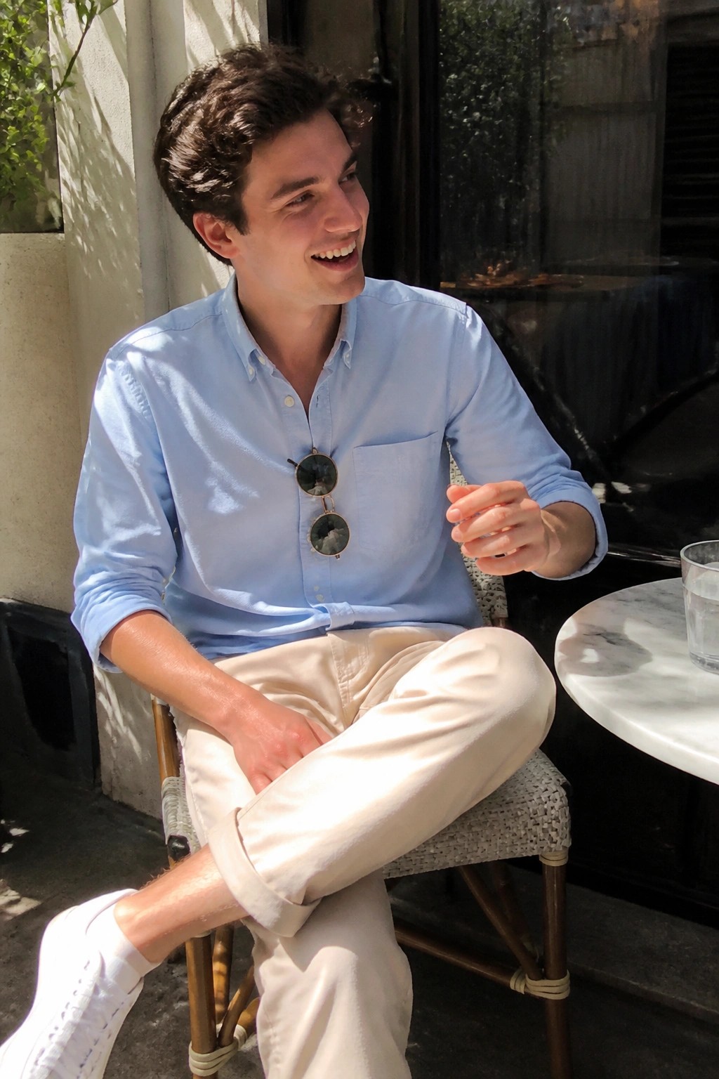 Young man in light blue shirt and cream pants smiling at outdoor table.
