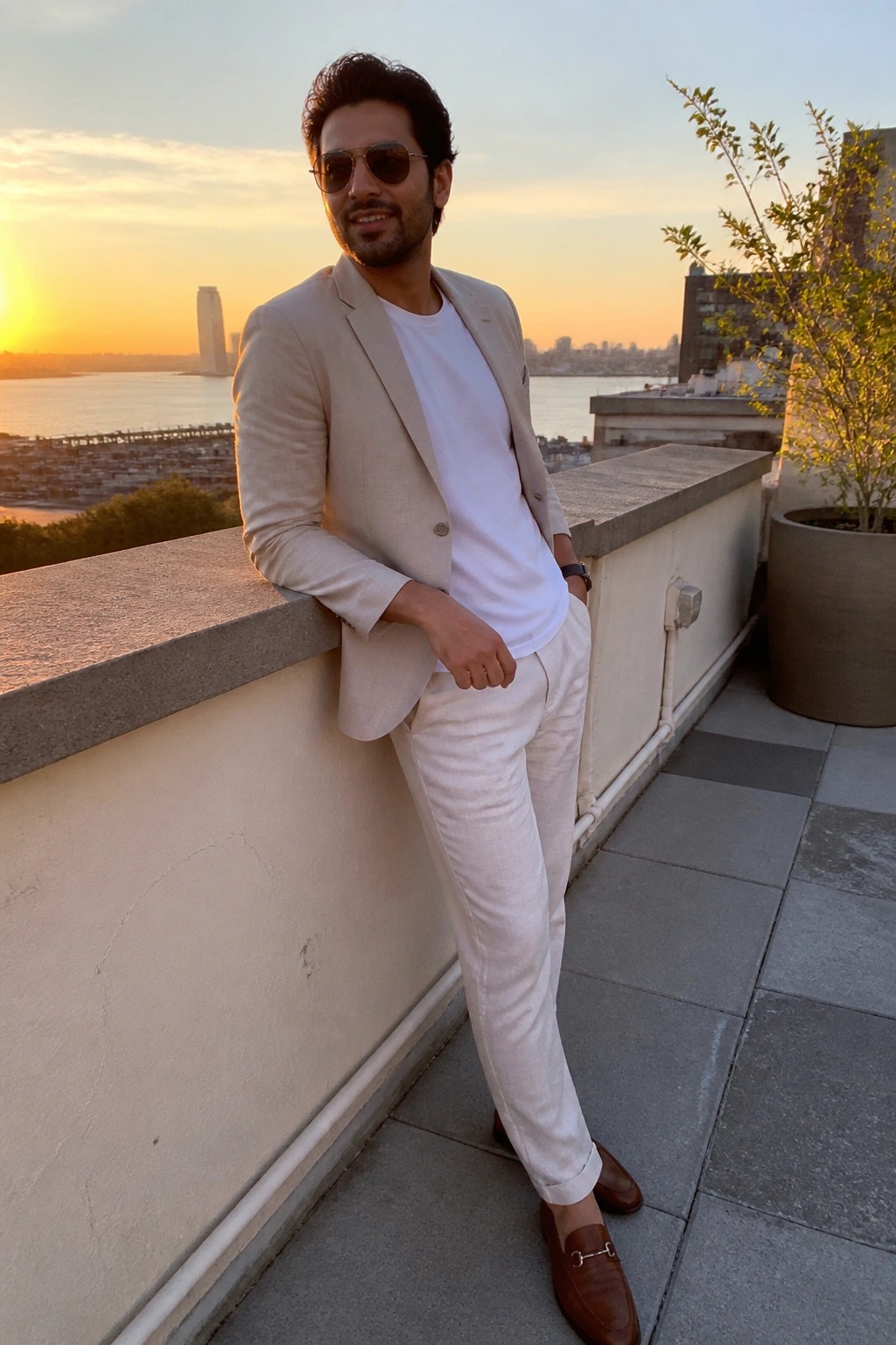 Man in beige blazer and white pants leaning on rooftop ledge at sunset