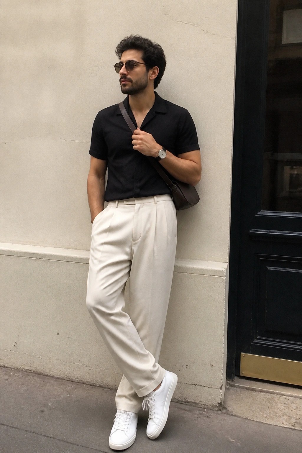 Man in black shirt and white pleated pants leaning against wall with bag