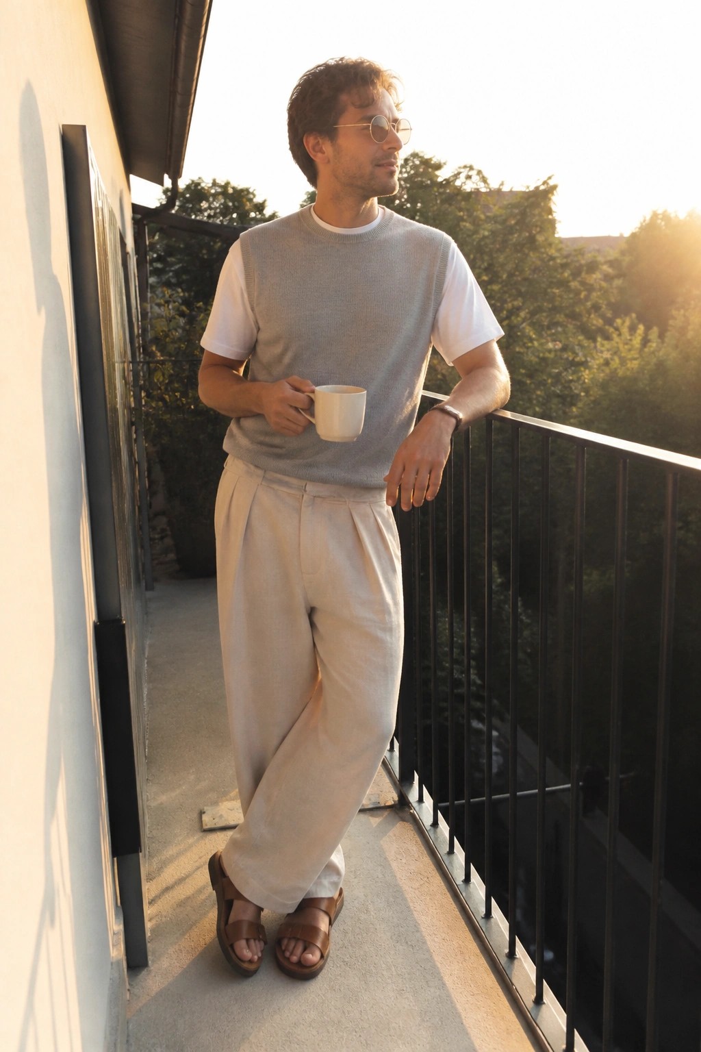 Man in gray sweater vest and beige pants holding mug on sunny balcony