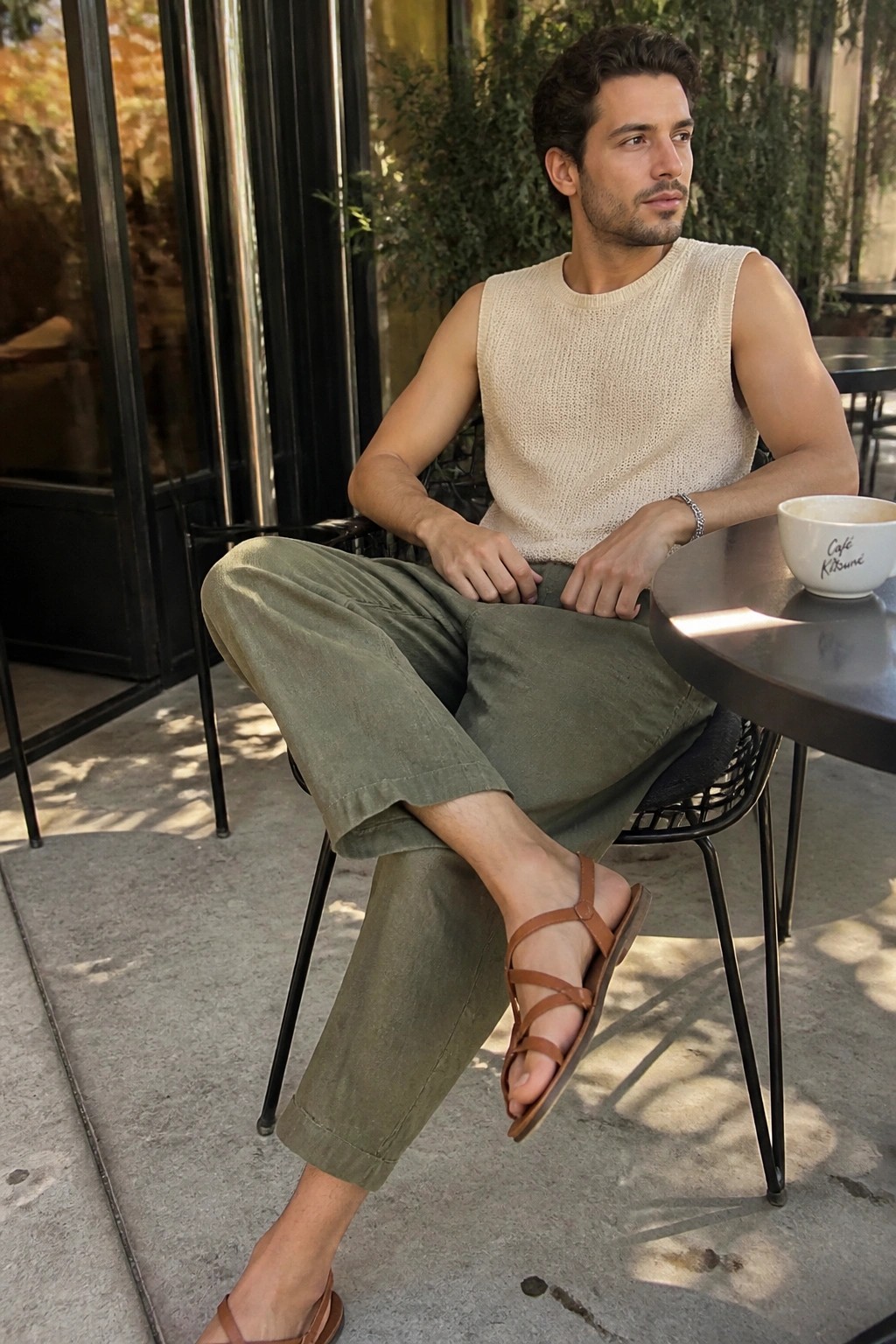 Man in beige sleeveless knit vest and olive pants at outdoor cafe table with sandals