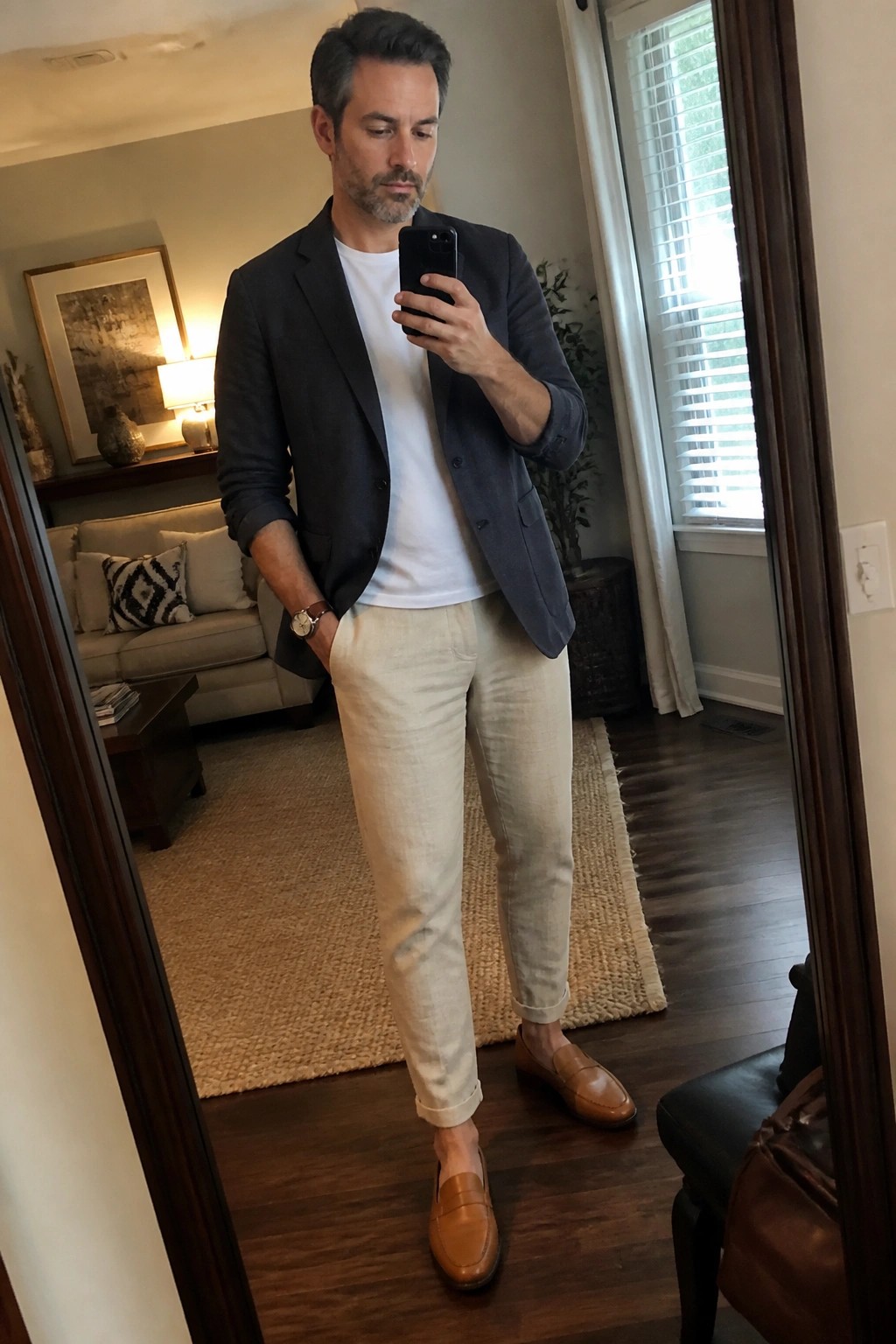 Man in dark blazer and beige pants taking mirror selfie in living room.