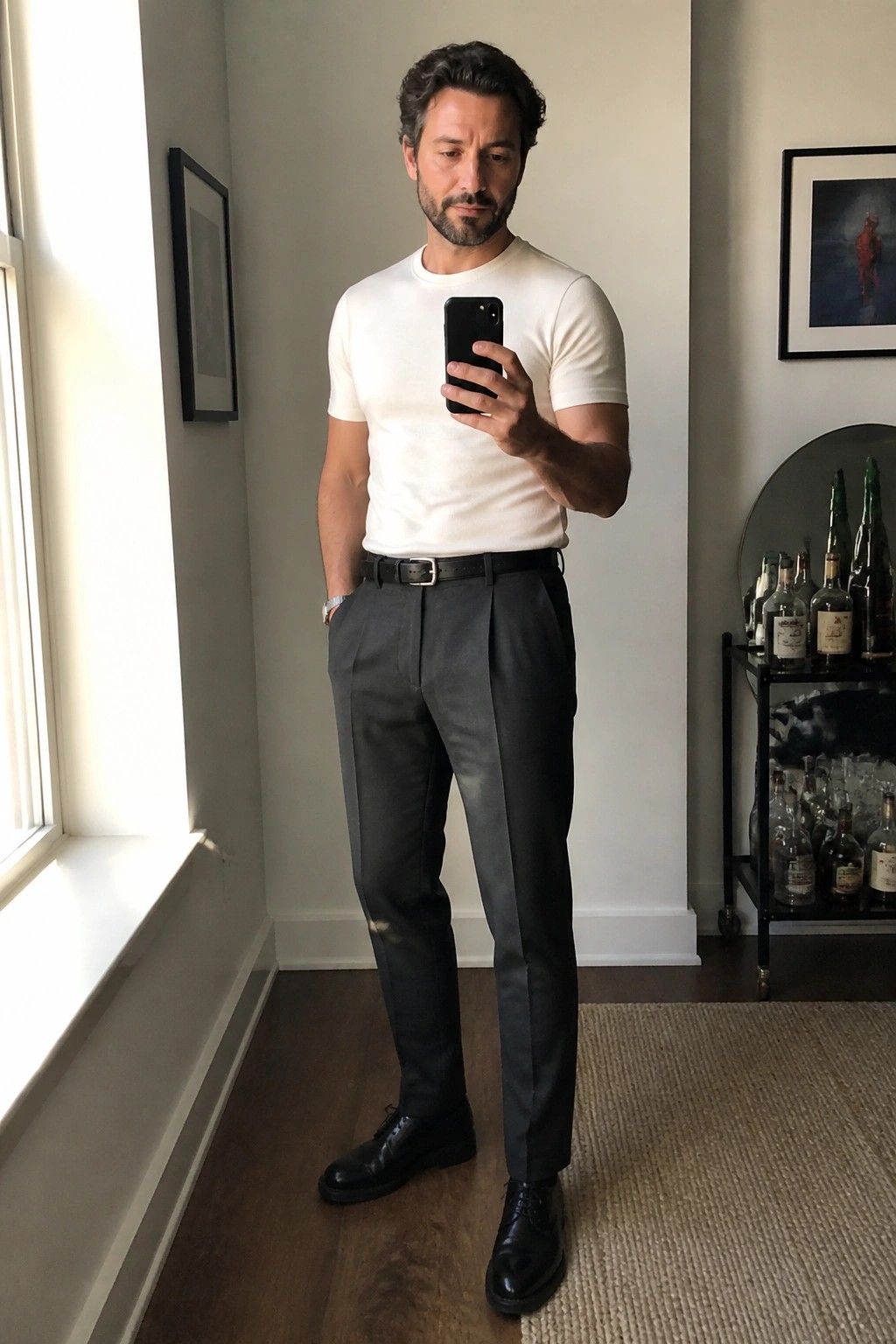 Man in white t-shirt and grey trousers taking mirror selfie indoors