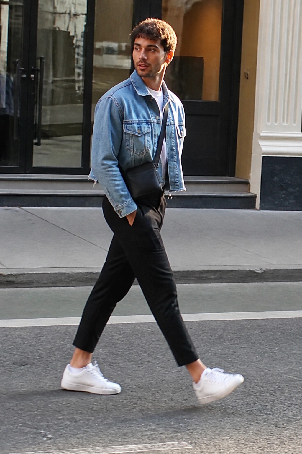 Man in denim jacket and black pants walking on city street with crossbody bag