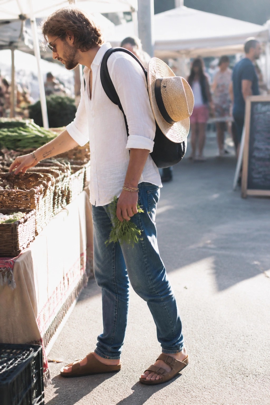 Man wearing a white linen button down shirt with rolled sleeves tucked into blue jeans, brown two strap sandals, black backpack holding a straw hat, standing at an outdoor market stall while holding fresh greens.