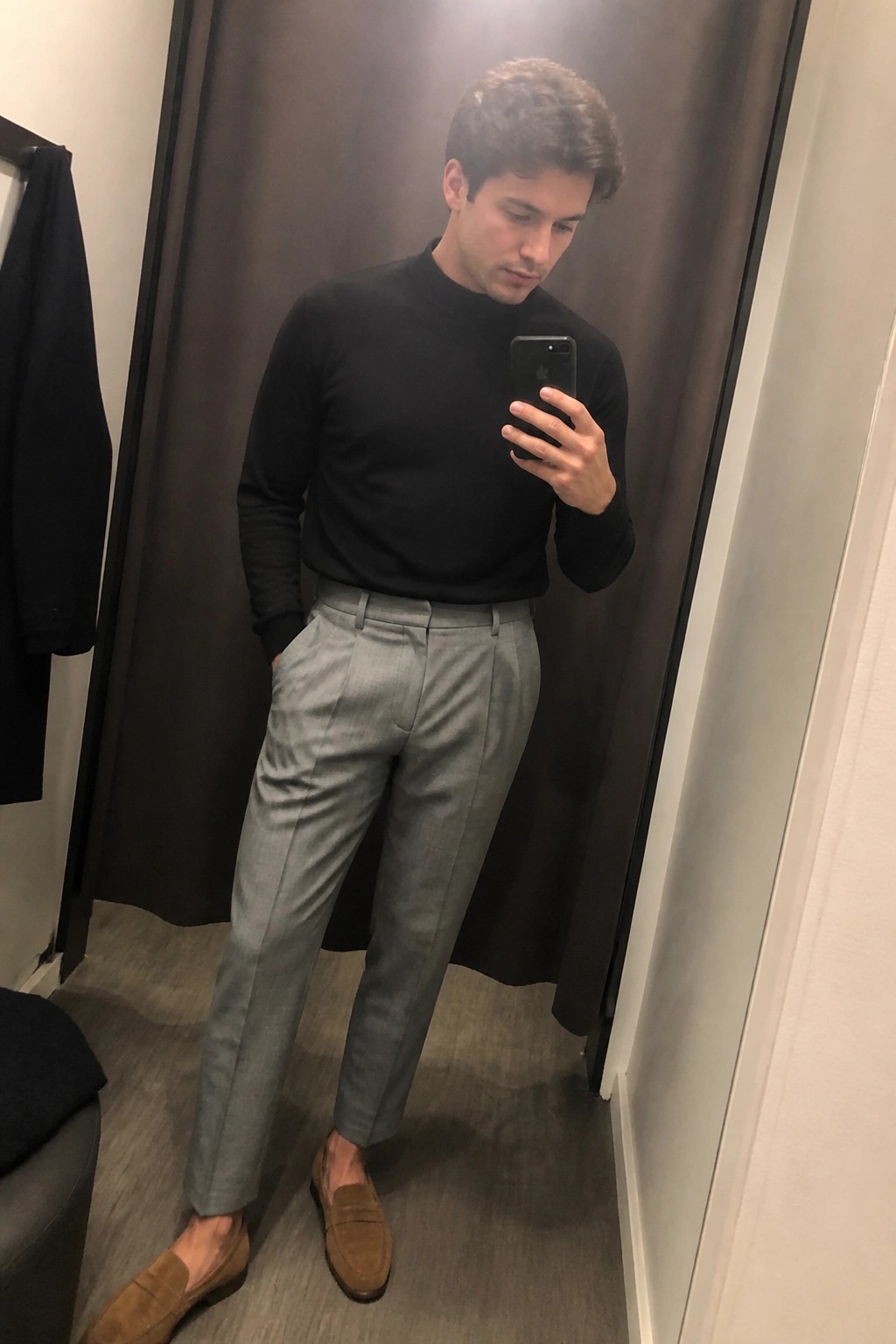 Man in black turtleneck, gray pleated trousers and brown suede loafers taking a mirror selfie indoors.