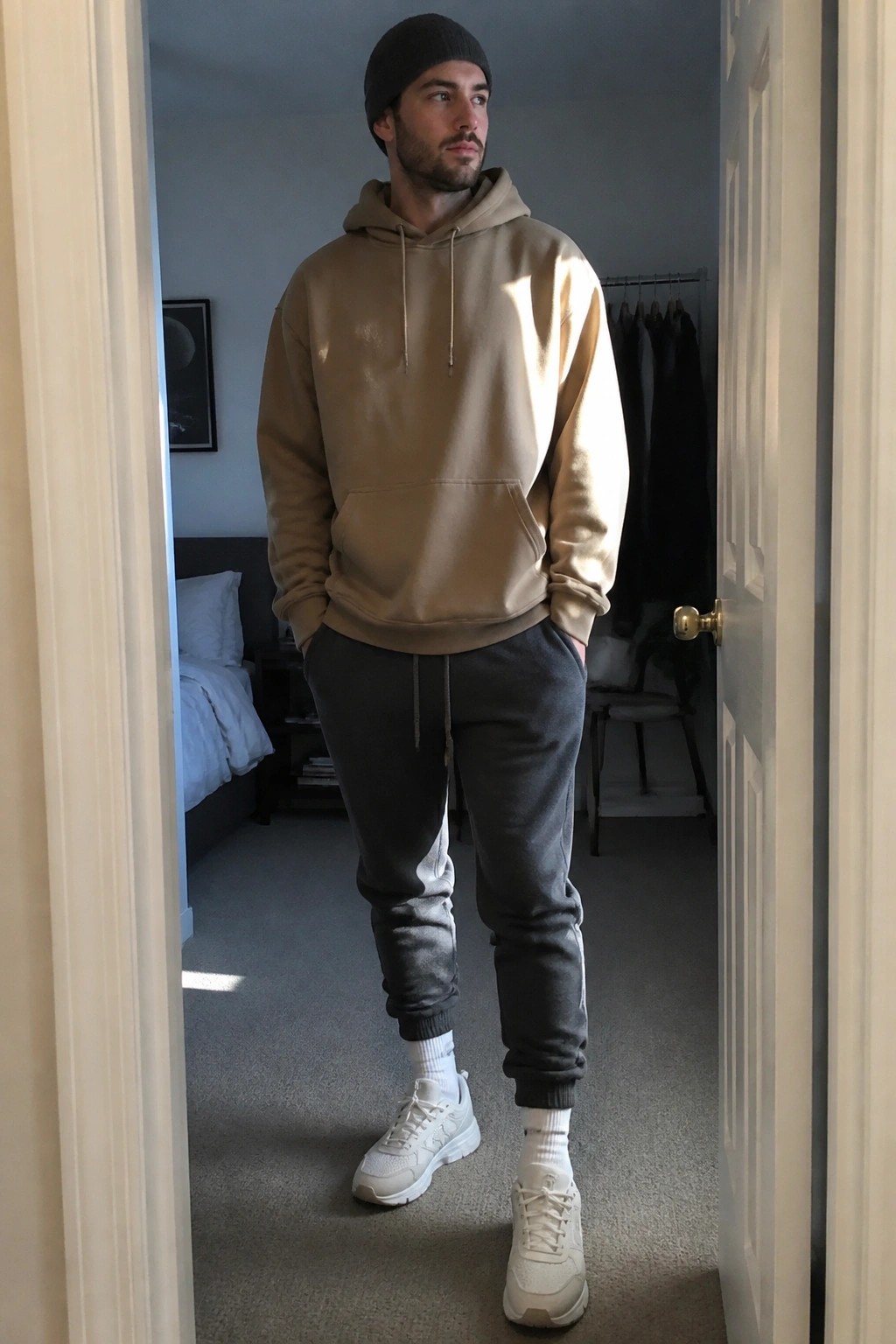 Man wearing beige hoodie, dark grey joggers with white side stripes, white sneakers, and black beanie in a bedroom doorway.