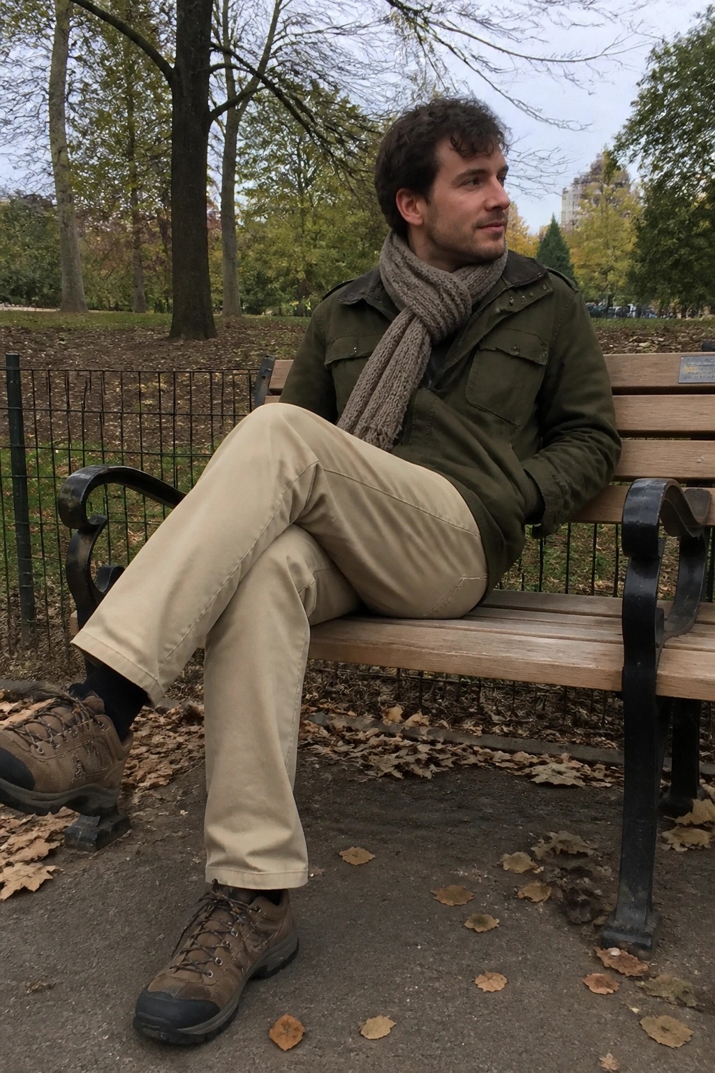 Man seated on park bench wearing olive green utility jacket, light gray knit scarf, beige khaki pants, and brown hiking boots.