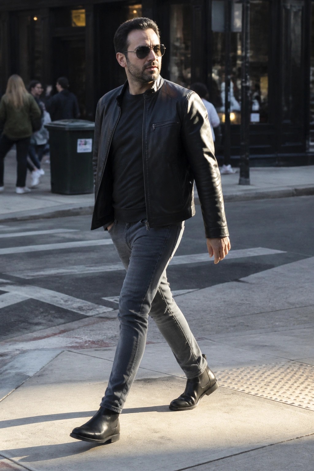Man in black leather jacket, gray jeans, and boots walking on sunny city street.