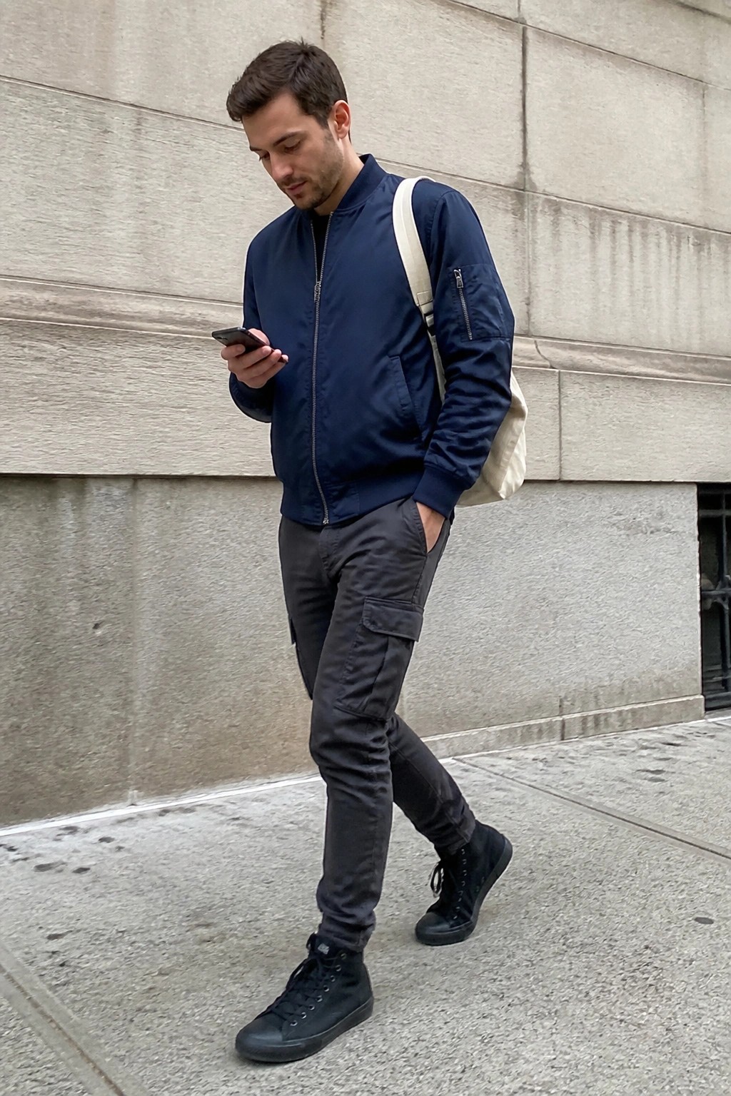 Man in navy bomber jacket checks phone while walking on sidewalk.