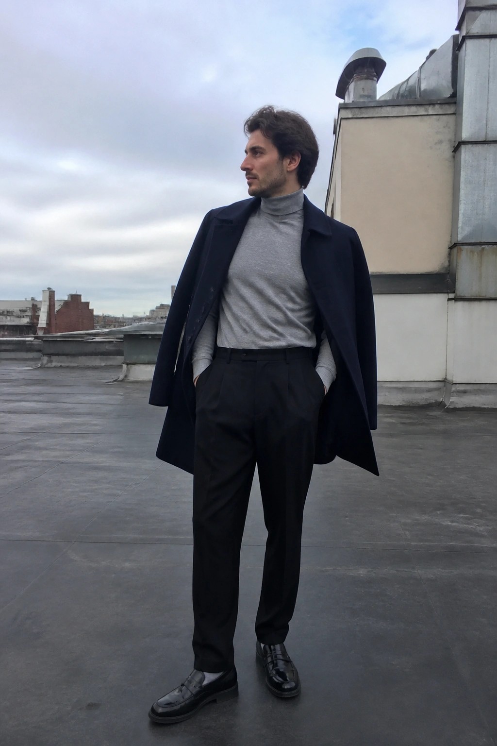Man in navy overcoat and gray turtleneck stands on rooftop, hands in pockets