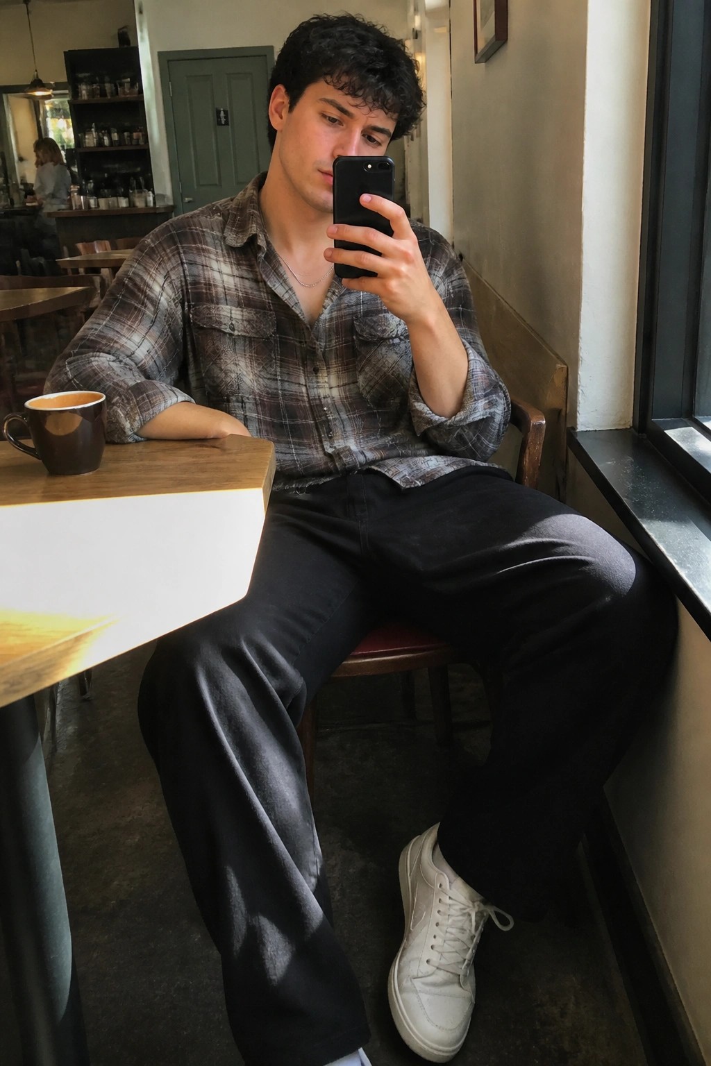 Young man in plaid shirt taking photo on phone in cafe with coffee mug.
