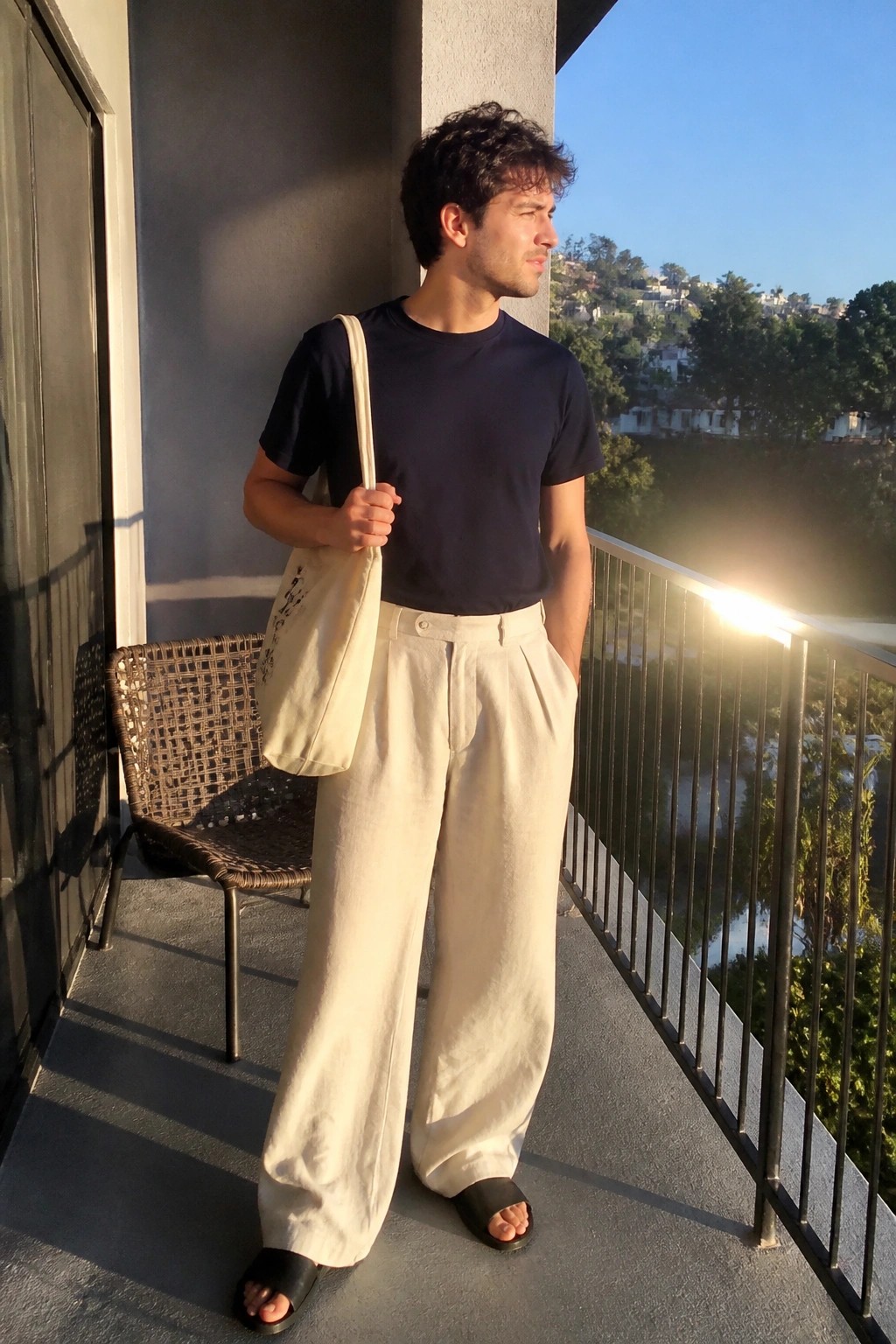 Man wearing a navy short sleeve tee, cream wide leg pleated pants, black slides, and carrying a canvas tote bag while standing on a balcony.