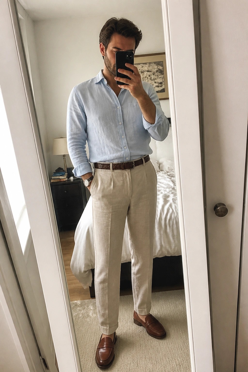 Man in light blue shirt and beige pants taking mirror selfie in bedroom