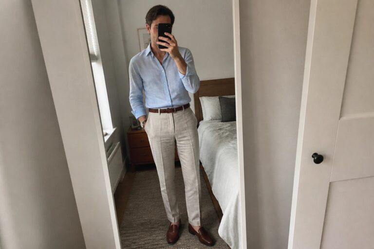 Man in light blue shirt and beige pants taking mirror selfie in bedroom