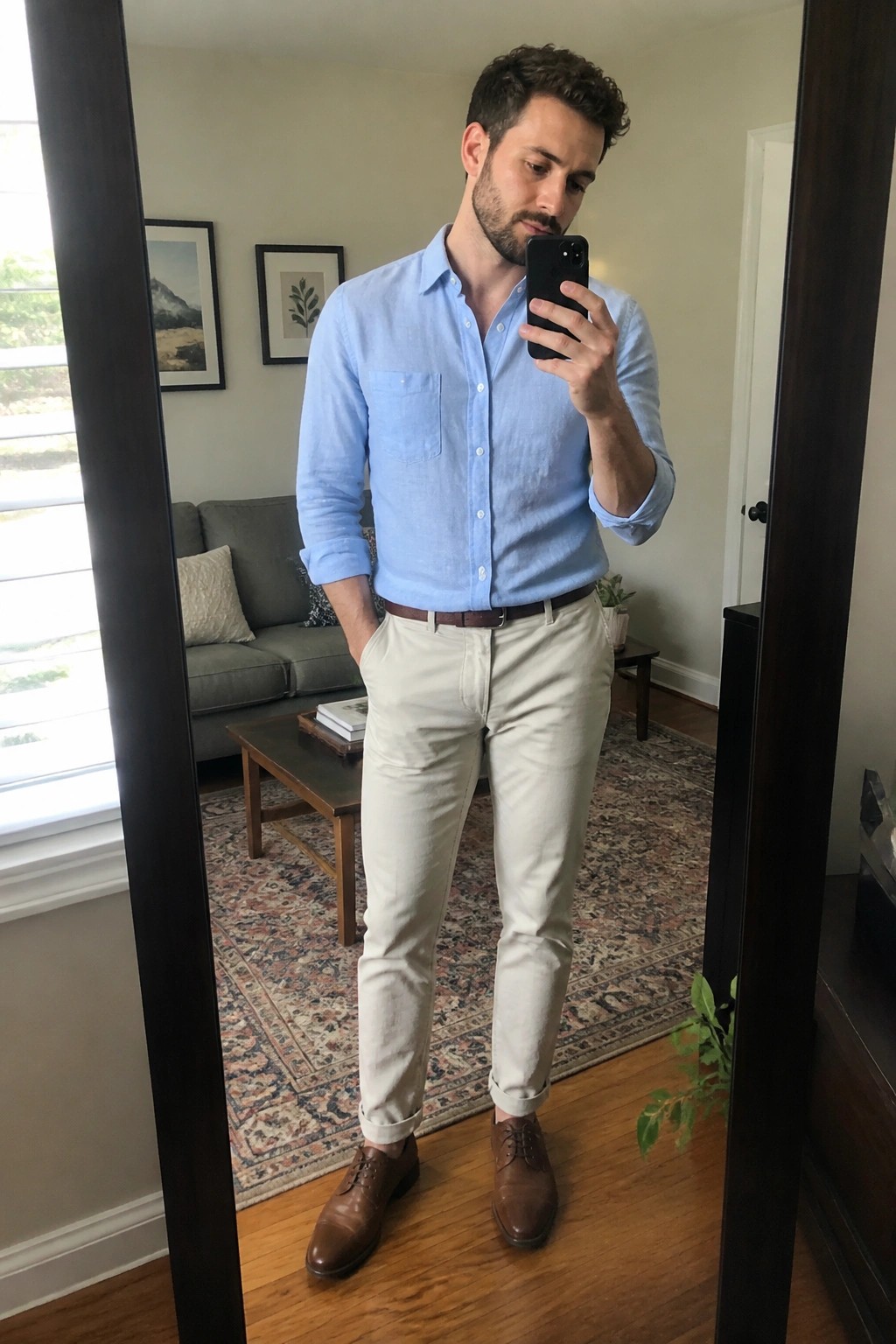 Man in light blue linen button down shirt with rolled sleeves, beige pants, brown belt and brown leather shoes taking a mirror selfie.