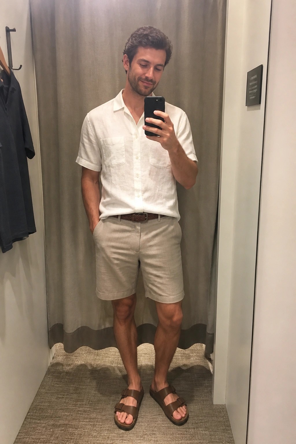 Man in white short sleeve linen button up shirt and beige linen shorts with brown belt and brown sandals taking a mirror selfie in a fitting room.