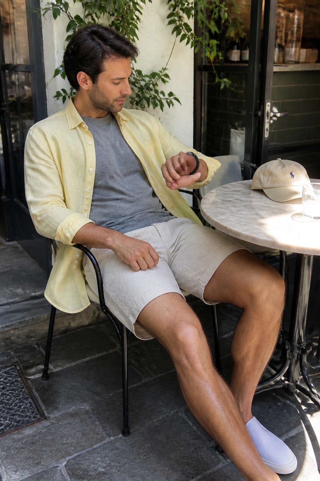 Man in yellow shirt checks watch at sunny outdoor cafe table with cap