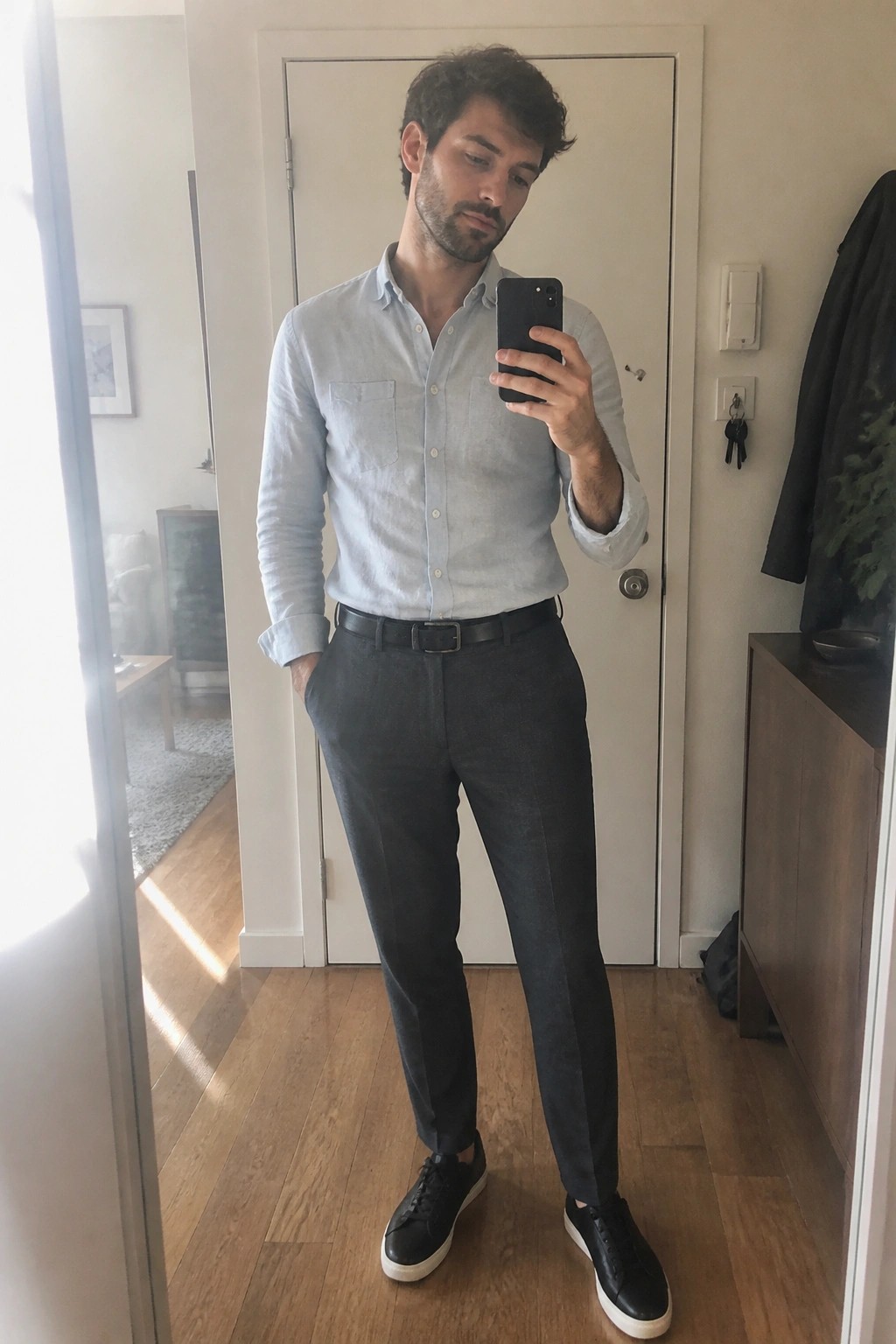 Man in light blue shirt and gray pants taking mirror selfie with phone.