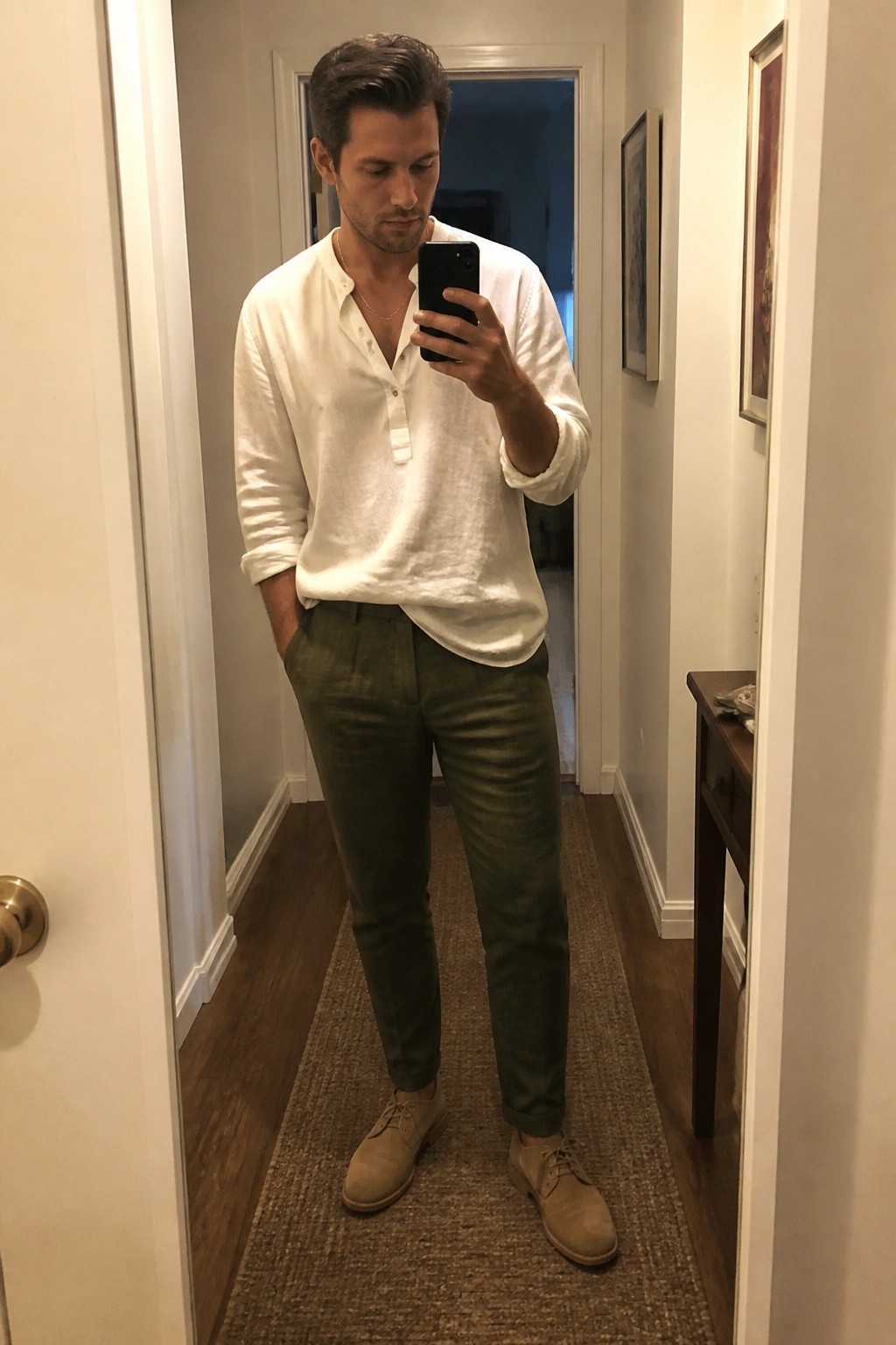 Man wearing a white linen long sleeve shirt tucked loosely into olive green linen pants with beige suede lace up shoes and a thin gold necklace while holding a phone in a hallway mirror selfie.