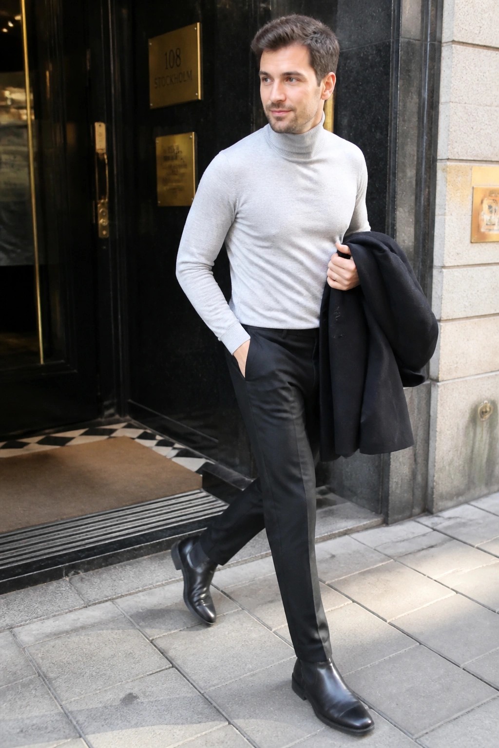 Man in gray turtleneck and black pants holding coat outside building entrance